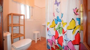 A bathroom with a toilet, a small stool, a shelf unit, and a shower curtain with a butterfly pattern. - Vrbo Property (Conil De La Frontera)