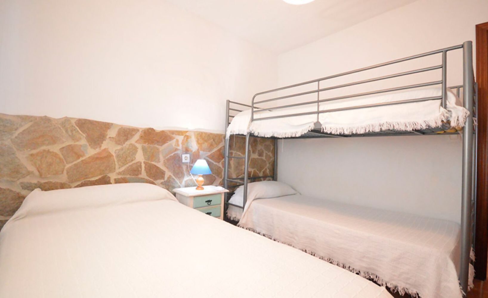 Habitación con literas, pared de piedra, mesita de noche con lámpara y puerta de madera.