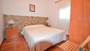 Un dormitorio con cama, dos mesitas de noche, pared de piedra, ventana con cortinas y una silla de madera.