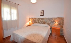 A bedroom with a bed, two bedside tables, a stone wall, a window with curtains, and a framed picture on the wall. - Vrbo Property (Conil De La Frontera)