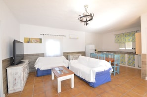 A room with two beds, a TV, a small table, a chandelier, and a kitchen area. - Samuel house with community pool and porch (Conil De La Frontera)