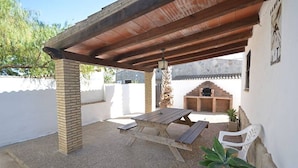 Een terras buiten met een houten tafel en banken, een barbecue van baksteen en een bloempot.