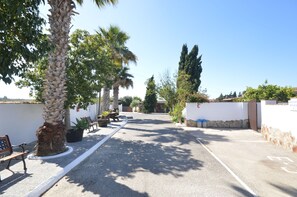A paved pathway lined with benches and potted plants, leading to a building with a stone wall and a clear blue sky.