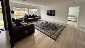 A spacious living room with a large window, a flat-screen TV, and a dark-colored sofa. - Beautiful 4 bedroom for short or long term stay! (Hastings)