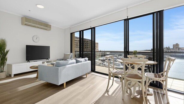 Un salon moderne avec un canapé, un téléviseur et un coin repas comprenant des chaises, de grandes fenêtres et une vue sur l’eau.