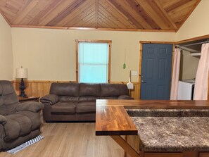 A living room with a brown leather sofa, a wooden coffee table, a lamp, a window with blinds, a blue door, and a washing machine.