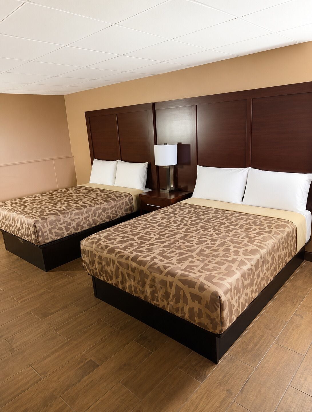 A hotel room with two beds, a wooden headboard, a nightstand with a lamp, and a wooden floor.