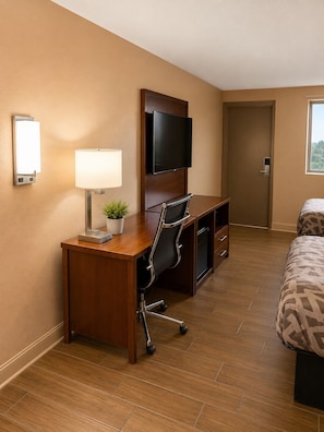 Ein Hotelzimmer mit einem Holztisch, einem Stuhl, einem an der Wand montierten Fernseher, einem Nachttisch, einem Bett und einem Fenster.