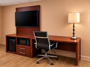 A hotel room with a wooden desk, a black office chair, a flat-screen TV mounted on the wall, a microwave, a lamp, and a built-in mini-fridge.