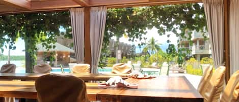 Un comedor luminoso con una mesa grande y sillas, ventanas amplias con cortinas, y vistas a una piscina y follaje.