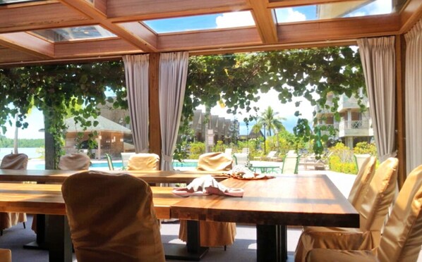 Un comedor luminoso con una mesa grande y sillas, ventanas amplias con cortinas, y vistas a una piscina y follaje.
