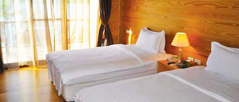 Habitación de hotel con dos camas, cabecero de madera, mesita de noche con lámpara y ventana con cortinas.