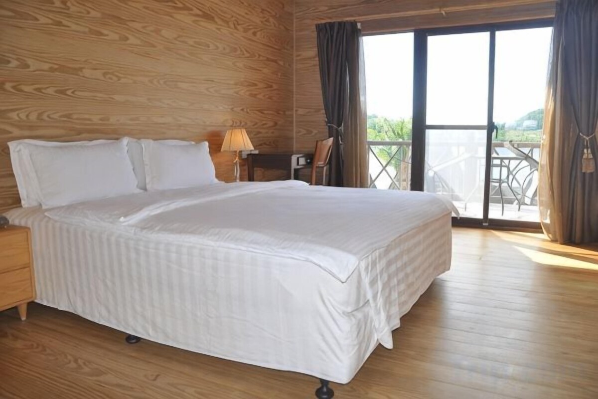 Un dormitorio con una cama grande, paredes de madera, una mesita de noche, una lámpara y vistas a un balcón.