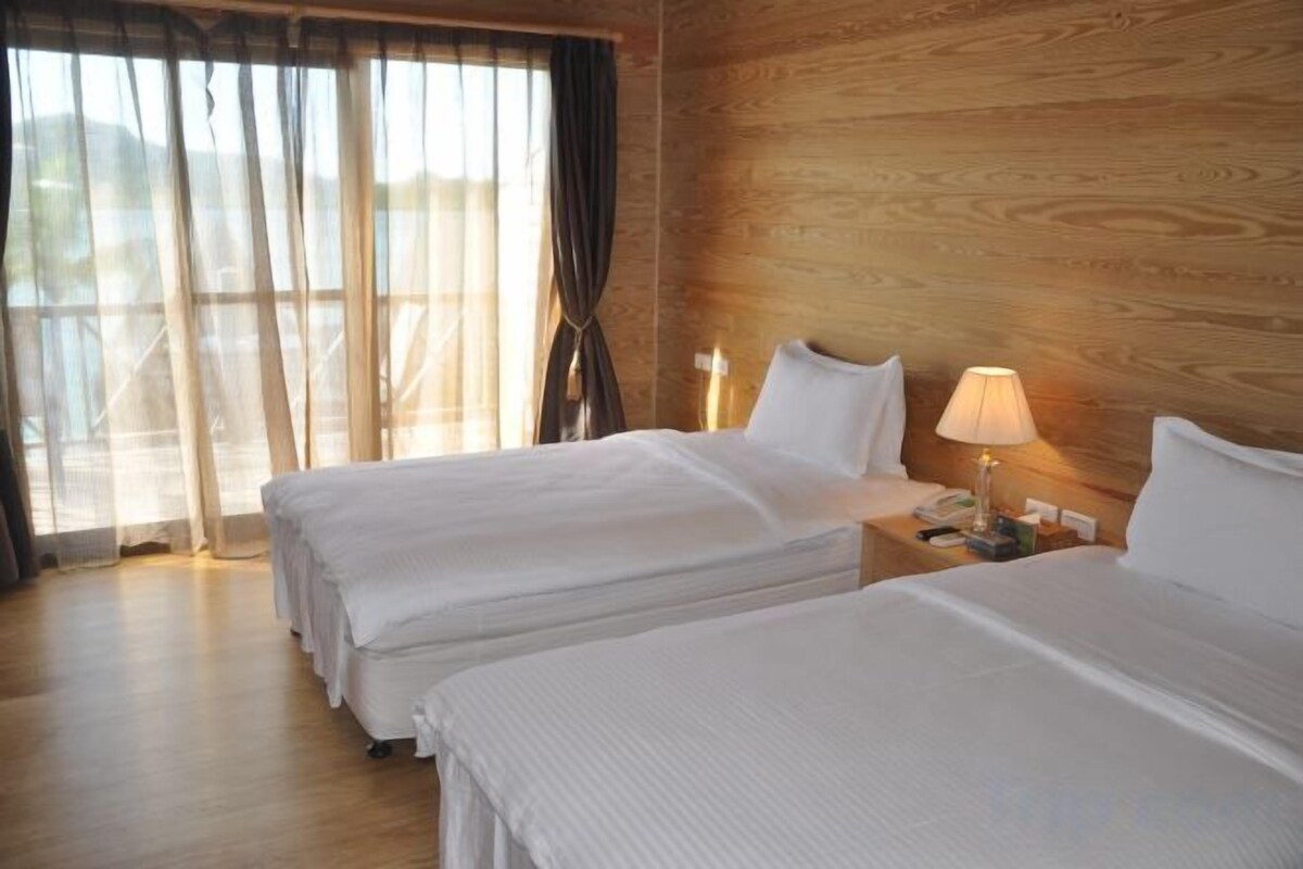 Habitación de hotel con dos camas, paredes de madera, una mesita de noche con lámpara y una ventana con cortinas.