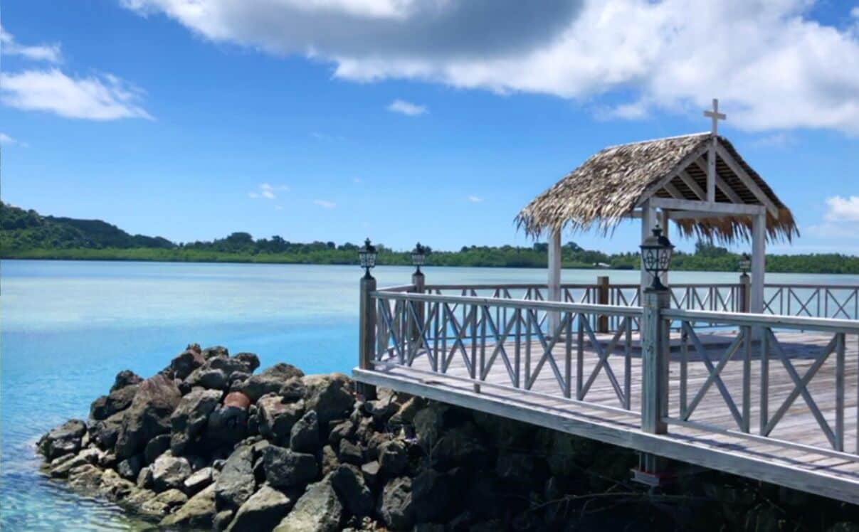 A pier with a thatched-roof structure, a railing, and a view of calm water and greenery.