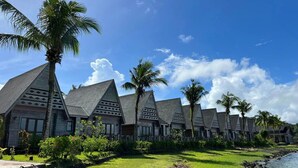 Une rangée de bâtiments de style bungalow, dotés de toits inclinés, entourés d’une végétation luxuriante et de palmiers, et situés à proximité d’une étendue d’eau.