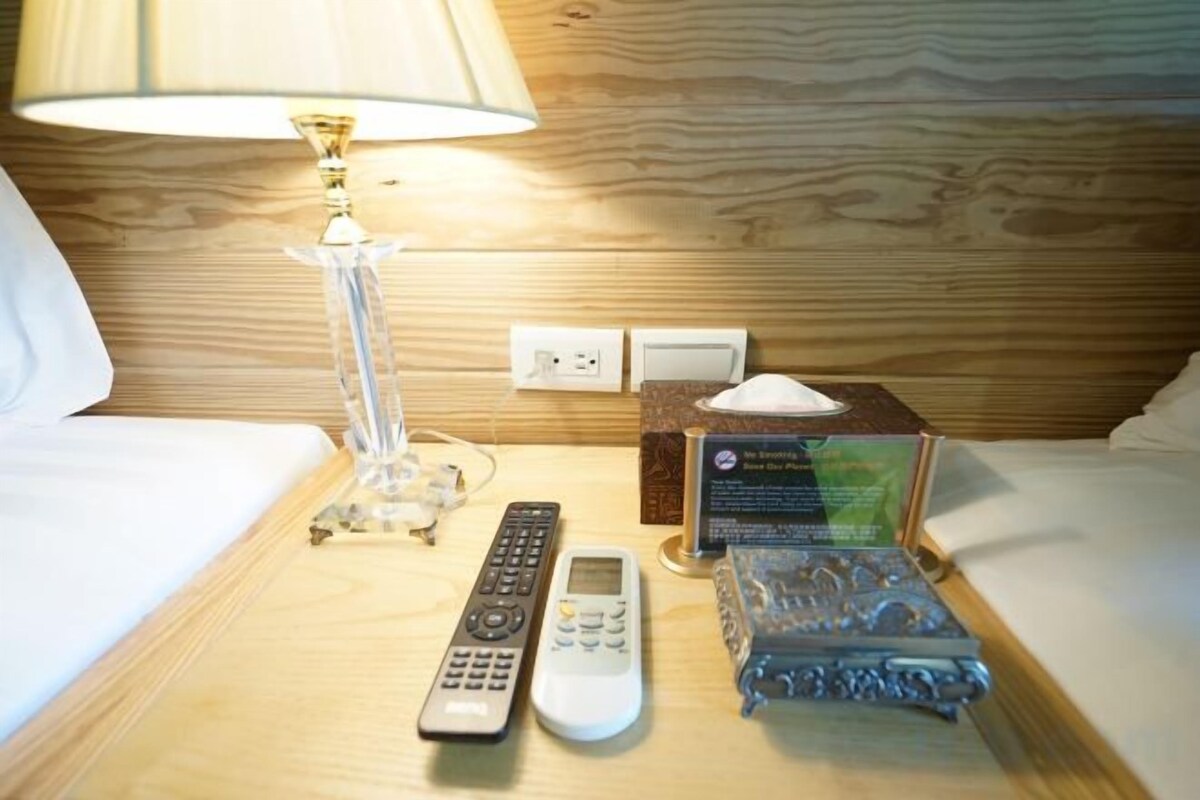 A neatly made bed with a white comforter, a bedside table with a lamp, a remote control, a mobile phone, and a decorative item.