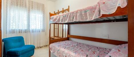 Una habitación con literas, una silla azul y una ventana con cortinas translúcidas.