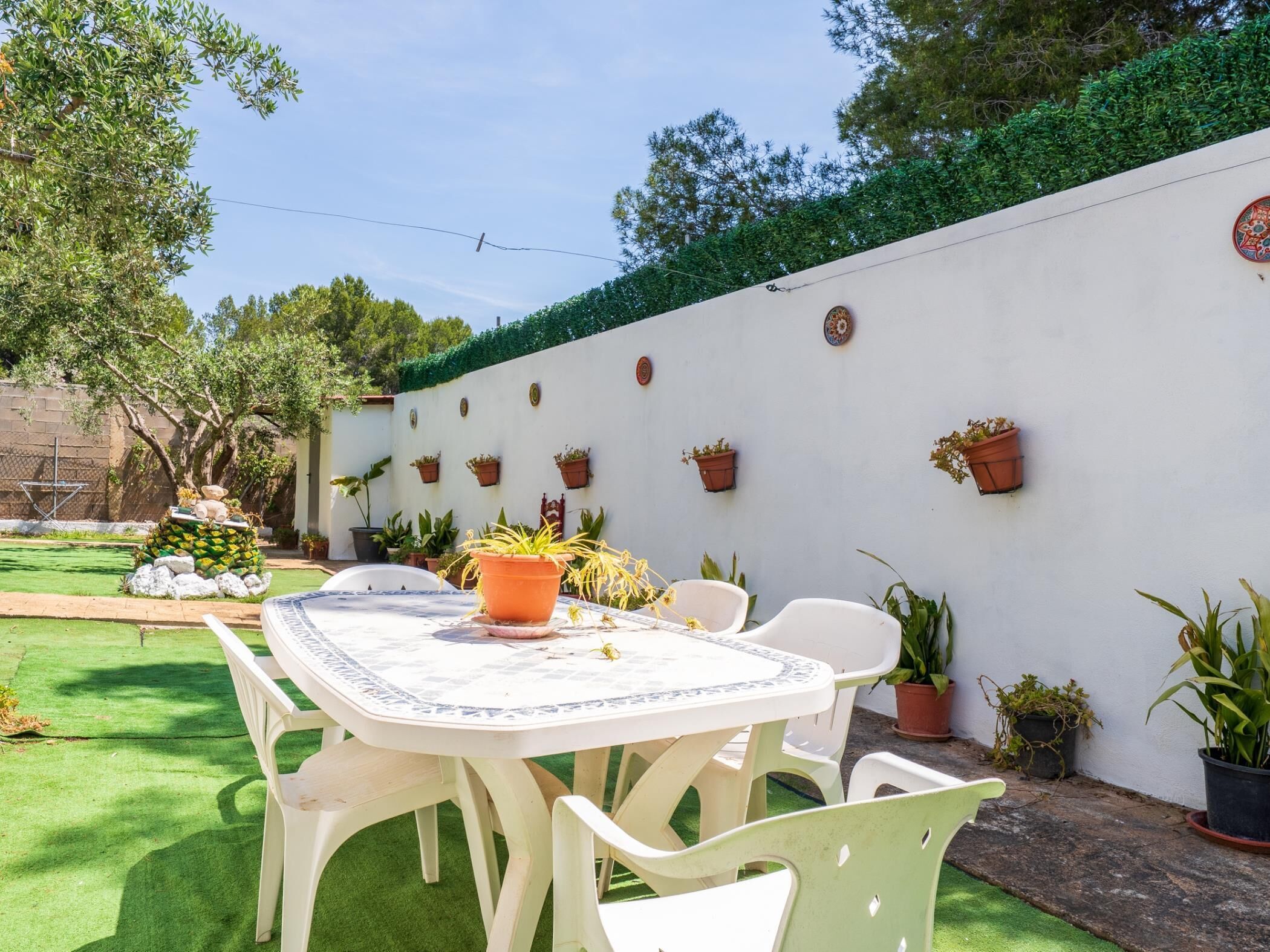 Eine Terrasse mit Tisch und Stühlen, umgeben von Kübelpflanzen und einer weißen Wand mit dekorativen Tellern.