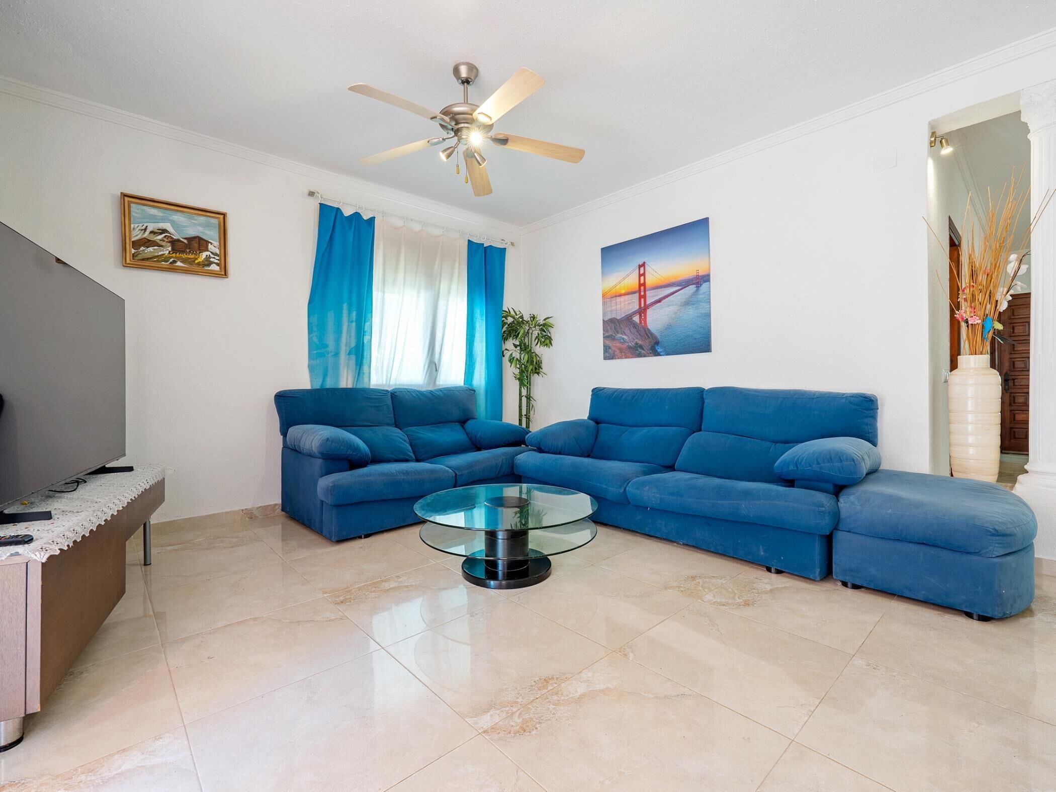 A living room with a sectional sofa, a glass coffee table, a ceiling fan, and a painting of a bridge.