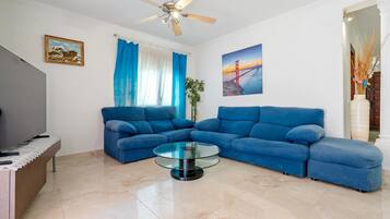 A living room with a sectional sofa, a glass coffee table, a ceiling fan, and a painting of a bridge.