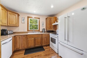 A kitchen with wooden cabinets, a white refrigerator, a stove, a sink, and a window with a view of greenery. - Walnut Grove- fenced yard- rural getaway (Coloma)