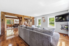 A spacious living room with a large gray sectional sofa, wooden beams, a mounted deer head, and a TV on the wall. - Walnut Grove- fenced yard- rural getaway (Coloma)
