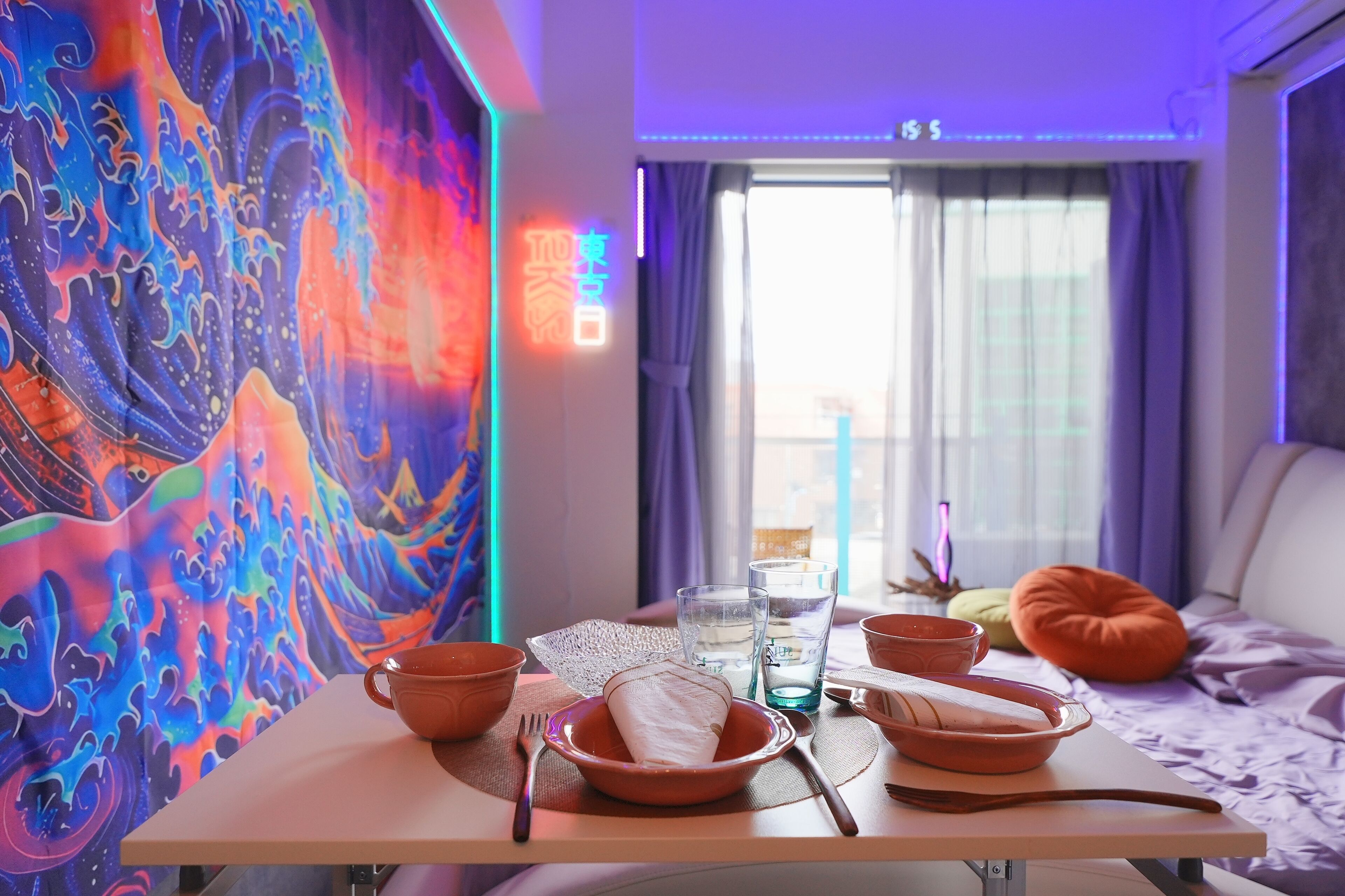 A dining area with a table set for two, a colorful tapestry on the wall, a bed with a pillow, and a window with curtains.