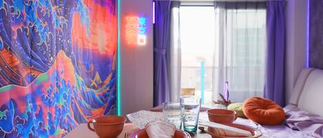 A dining area with a table set for two, a colorful tapestry on the wall, a bed with a pillow, and a window with curtains.