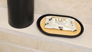Ett svart, cylindriskt föremål och en bricka med ett paket på, där det står ”LUX BOTANICALS JAZMIN CREMOSO” på etiketten. Allt detta ligger på en beige yta.
