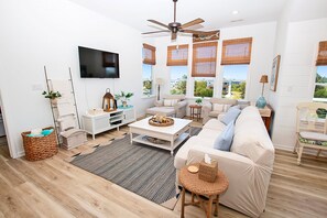 Ein modernes Wohnzimmer mit Deckenventilator, Flachbildfernseher, einer Couch, einem Couchtisch und Blick ins Freie.