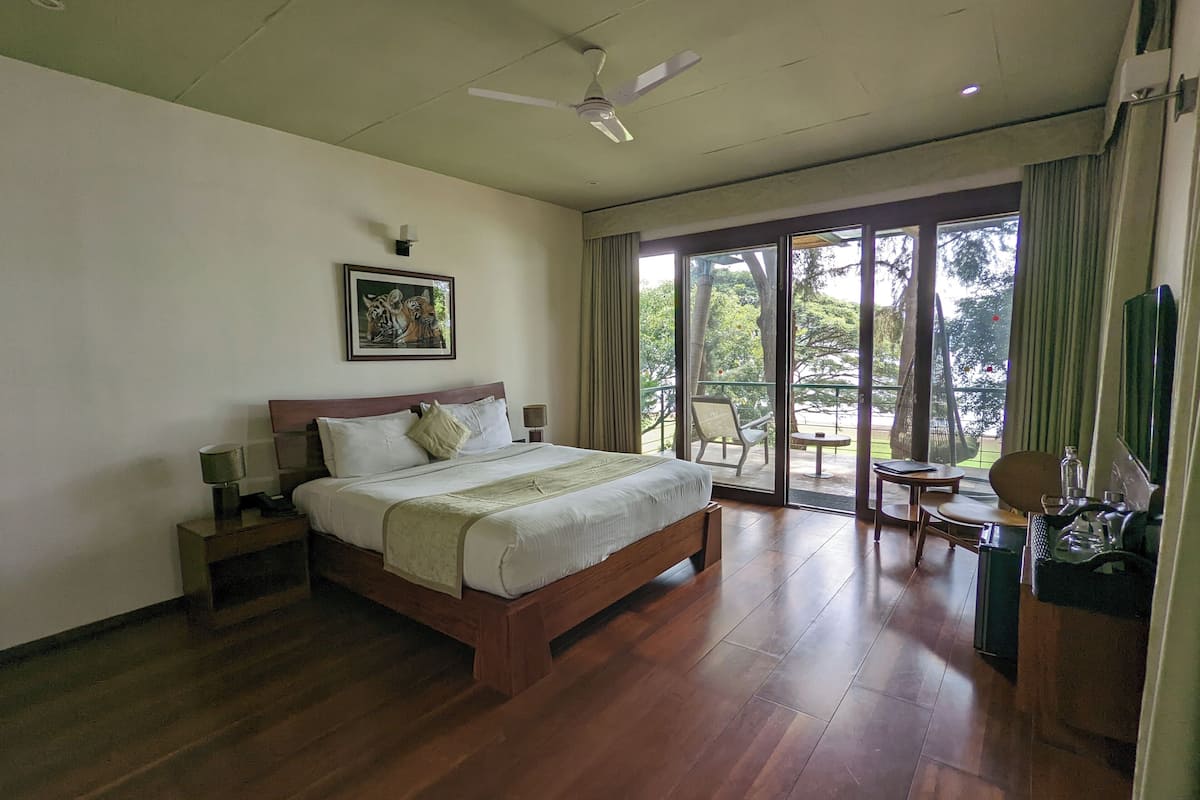 A hotel room with a wooden bed, a ceiling fan, a painting on the wall, and a view of the outdoors through a sliding door.