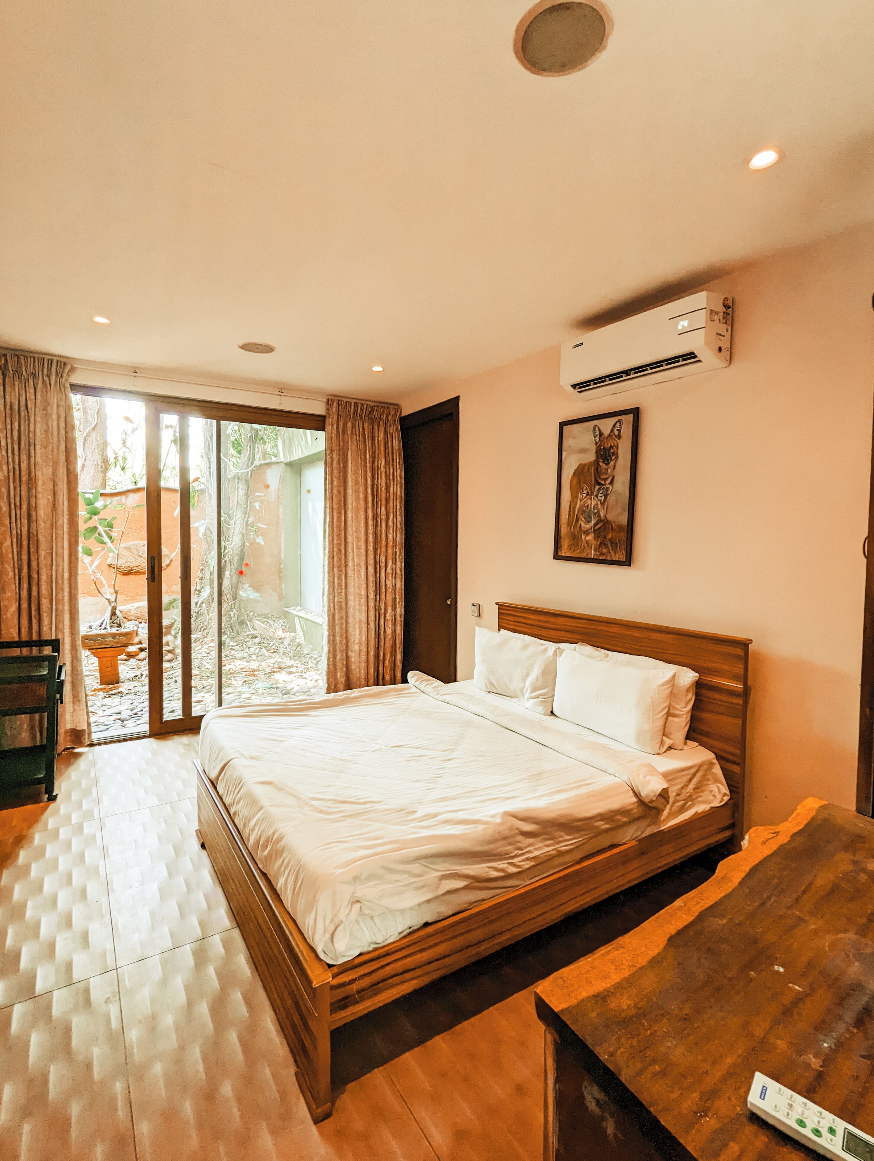 Une chambre à coucher avec un lit, une tête de lit en bois, un tableau, un appareil de climatisation et une vue sur l’extérieur, visible à travers une porte vitrée.