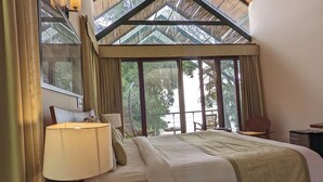 A bedroom with a large bed, a nightstand, a lamp, a chair, and a view of the outdoors through large windows. - Waterwoods Lodge Kabini (Heggadadevankote)