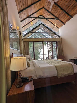 A bedroom with a large bed, a nightstand, a lamp, a chair, and a view of the outdoors through large windows. - Waterwoods Lodge Kabini (Heggadadevankote)
