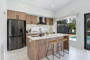 A modern kitchen with a central island, stainless steel appliances, wooden cabinetry, and a view of a swimming pool. - Belle Escapes The Trio In Trinity (Trinity Beach)
