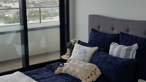Ein Schlafzimmer mit großem Fenster, einem Bett mit dunkelblauer Bettwäsche und Blick auf eine Stadtlandschaft.
