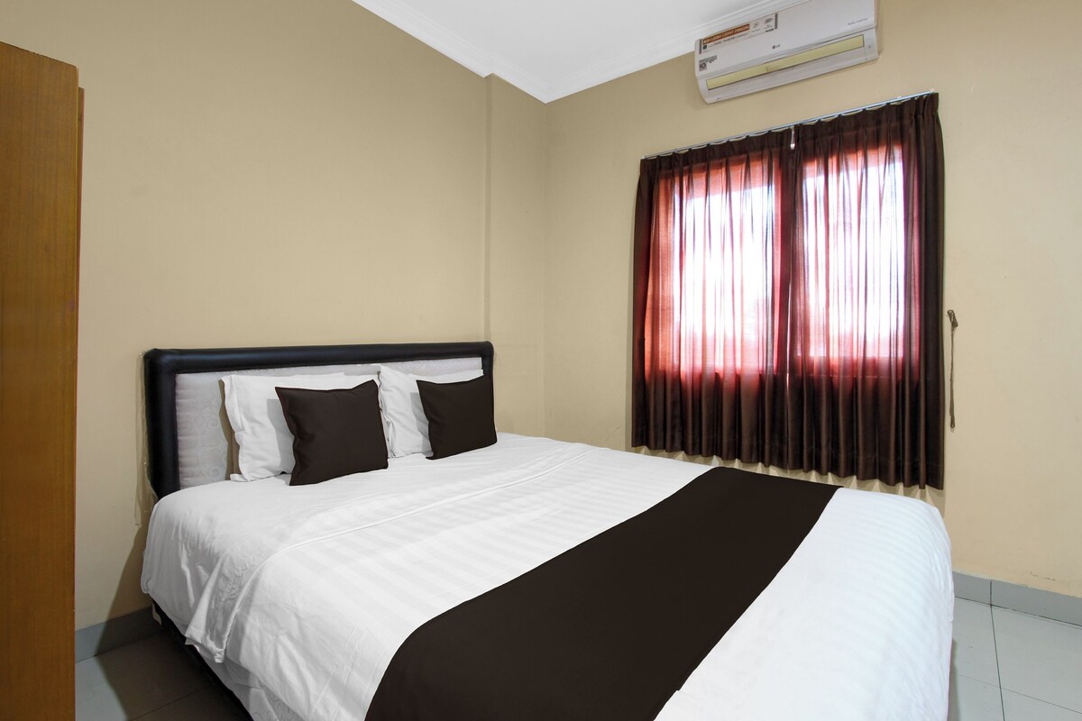A hotel room with a bed, a window with curtains, an air conditioning unit, and a door.