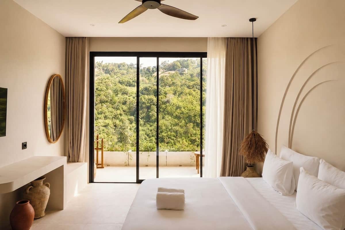 A bedroom with a large bed, a ceiling fan, a view of greenery through a sliding door, and a vase on the nightstand.
