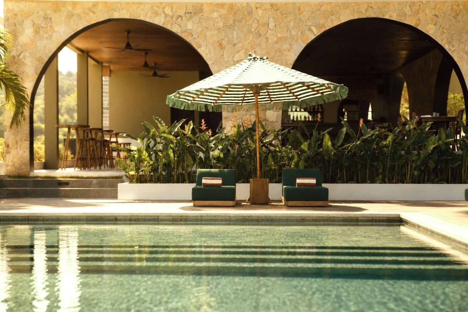 A pool area with two green sofas and a striped umbrella, surrounded by plants and stone arches.