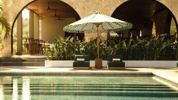 A pool area with two green sofas and a striped umbrella, surrounded by plants and stone arches.
