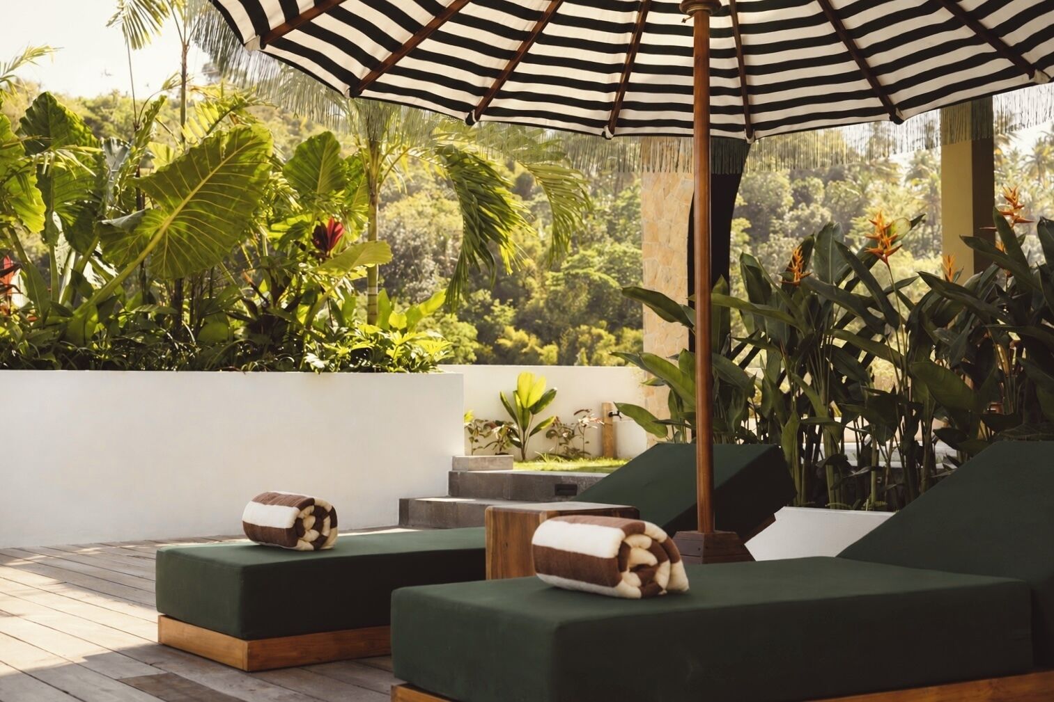 A tropical outdoor lounge area with green cushioned seating, a striped umbrella, and lush greenery in the background.