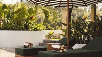 A tropical outdoor lounge area with green cushioned seating, a striped umbrella, and lush greenery in the background.
