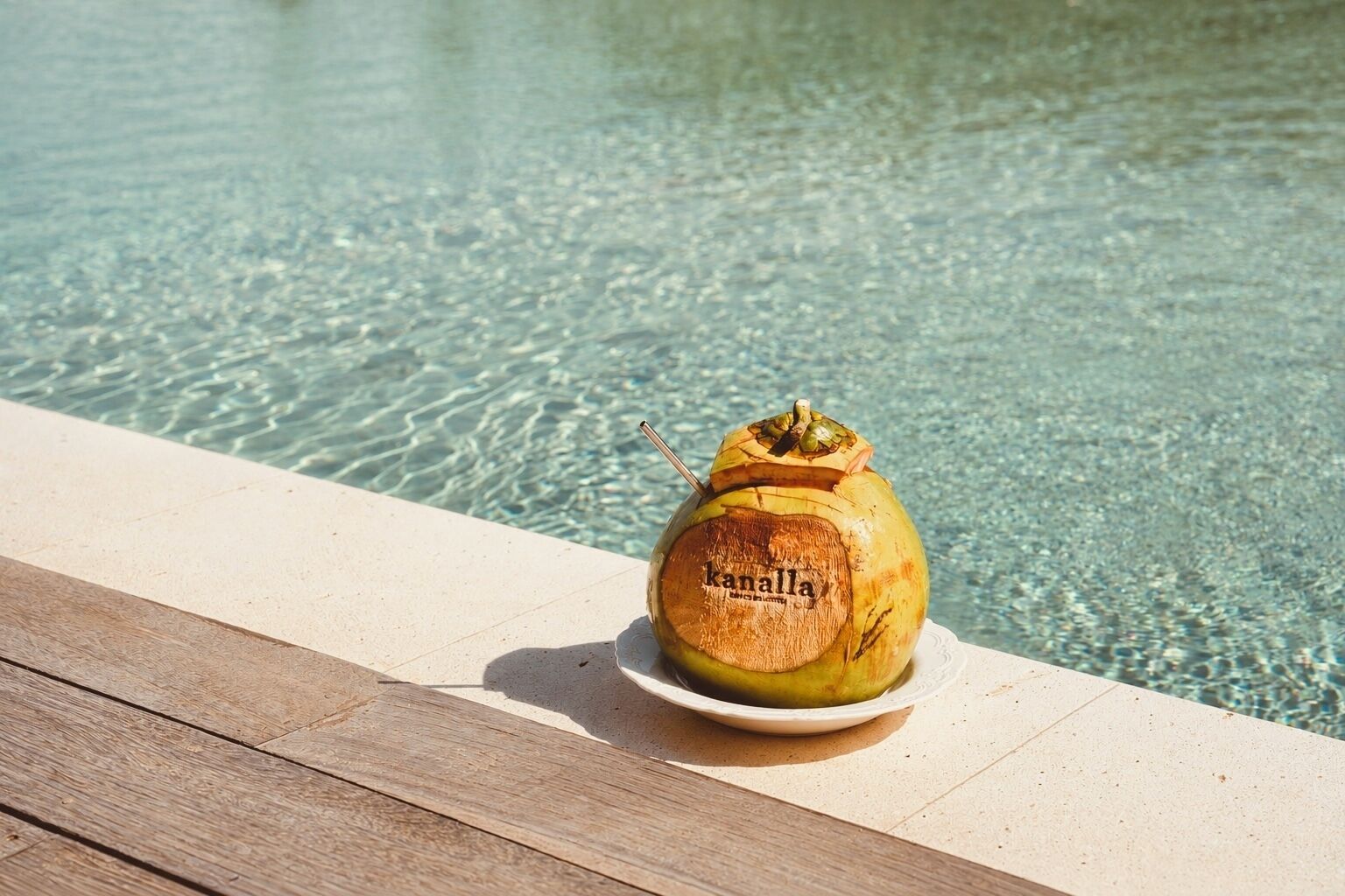 A coconut with a straw and the word 'kanalla' on a white plate by a pool.