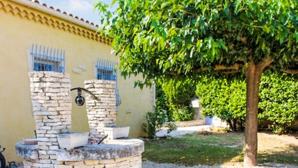 Una fuente de piedra con un recipiente metálico, una pared con rejas azules en las ventanas y un árbol al fondo.