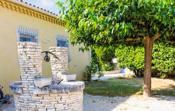 A stone fountain with a metal basin, a wall with blue window bars, and a tree in the background. - Maison piscine proche Alpilles (Senas)