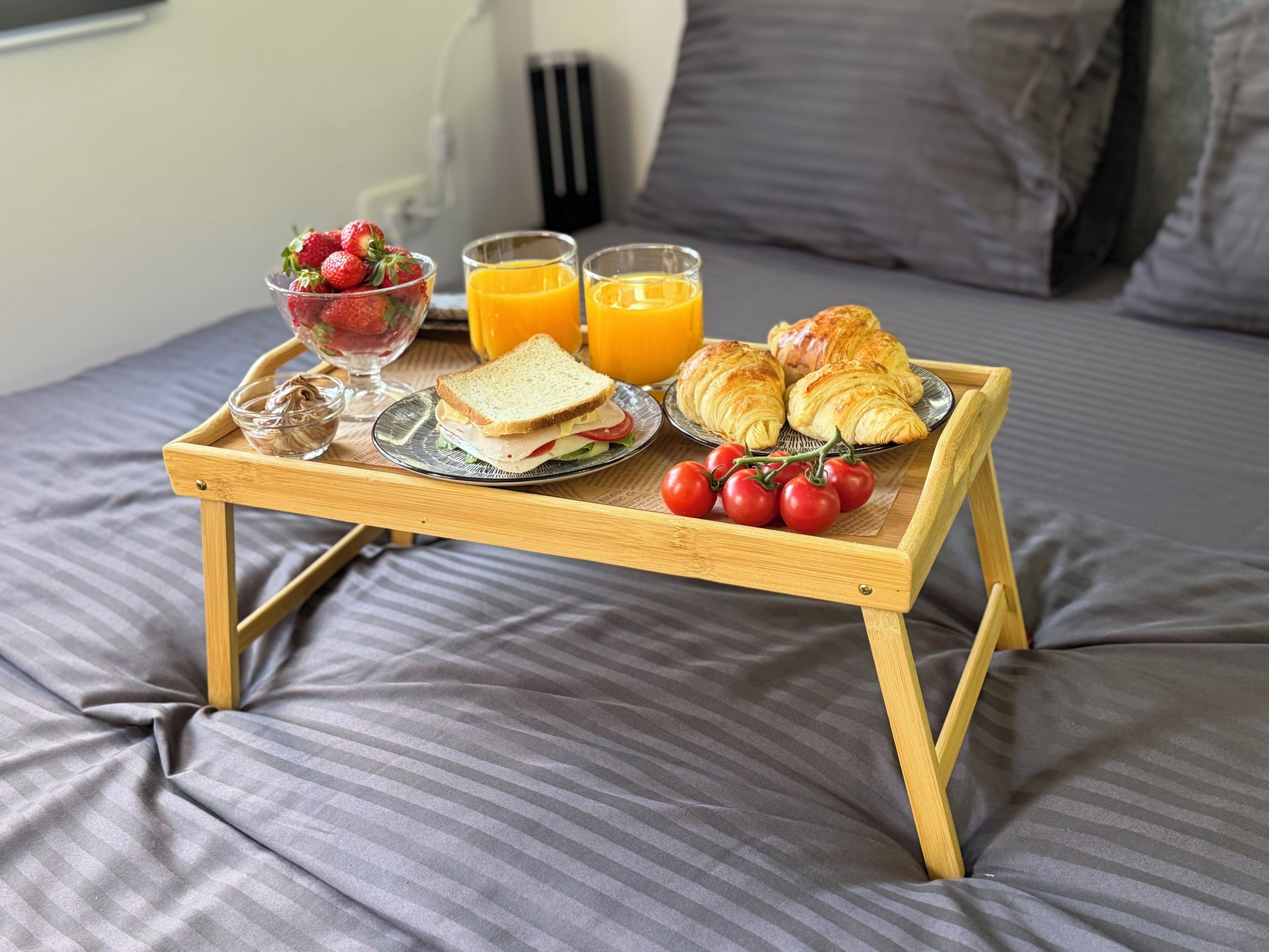 A breakfast tray on a bed with croissants, tomatoes, a sandwich, and orange juice.