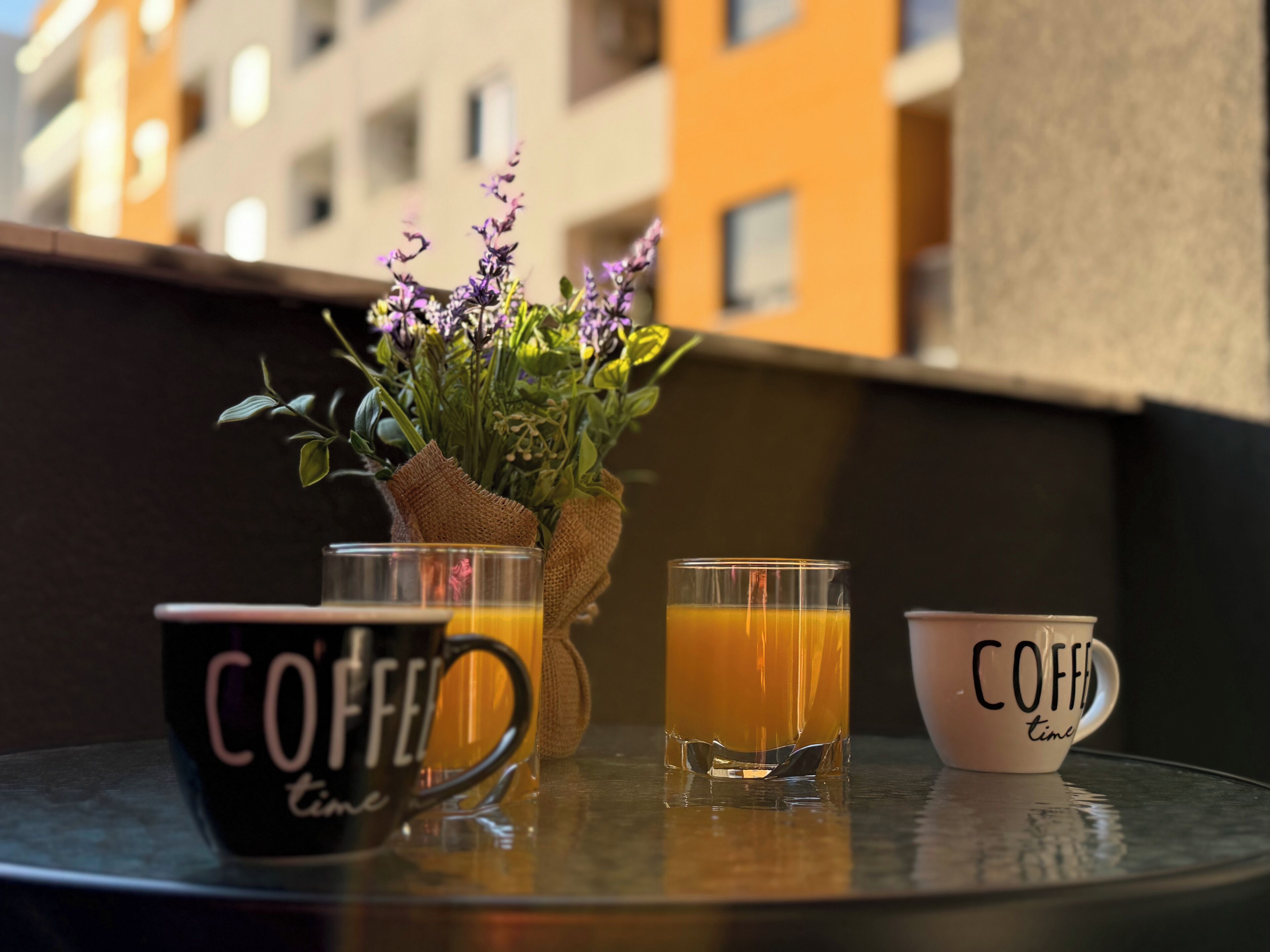 A table with two coffee mugs and two glasses of orange juice, a small plant, and a burlap sack.