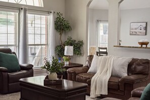 Ein Wohnzimmer mit braunem Ledersofa, einem Couchtisch und einem Kamin. Es gibt Fenster mit Jalousien, eine Pflanze und ein Gemälde an der Wand.