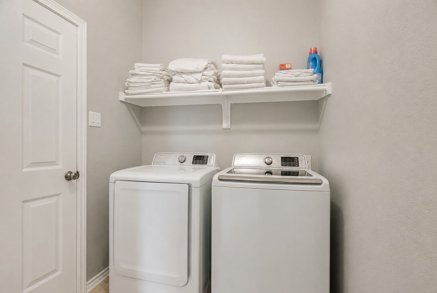 A laundry area with two white washers and dryers, a shelf with folded towels, and a door.